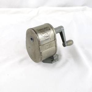 May include: Vintage silver-toned pencil sharpener with a hand crank. The sharpener has a cylindrical body and a base. The text "CHICAGO" is visible on the side. A classic office or school supply.