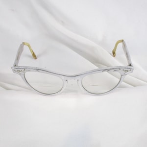 May include: Vintage cat-eye eyeglasses with a silver frame and clear lenses. The temples are a light yellow color. The glasses are set against a white background.