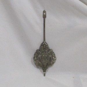 May include: A silver-toned clock pendulum with an ornate, floral-inspired design. The pendulum has a long, slender stem with a loop at the top and a decorative, shield-shaped weight at the bottom. The weight features a central emblem.