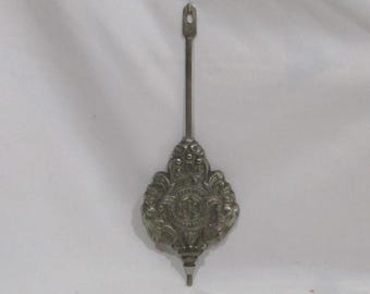 May include: A silver-toned clock pendulum with an ornate, floral-inspired design. The pendulum has a long, slender stem with a loop at the top and a decorative, shield-shaped weight at the bottom. The weight features a central emblem.