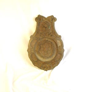 May include: Ornate, antique brass wall plaque with a detailed floral and scroll design. The plaque has a central circular medallion with a floral pattern and an elaborate, decorative border. The overall shape is elongated with a curved top.