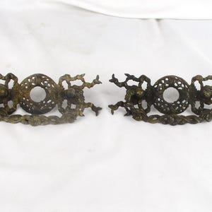 May include: Two antique bronze drawer pulls with intricate, ornate designs. Each pull features a central circular element with a decorative pattern, flanked by detailed, branch-like embellishments. The pulls are set against a white background.