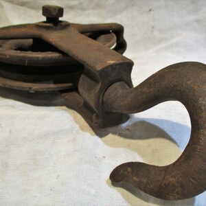 May include: A rusty cast iron pulley with a hook. The pulley has two wheels and a handle. The hook is bent at a 90-degree angle.