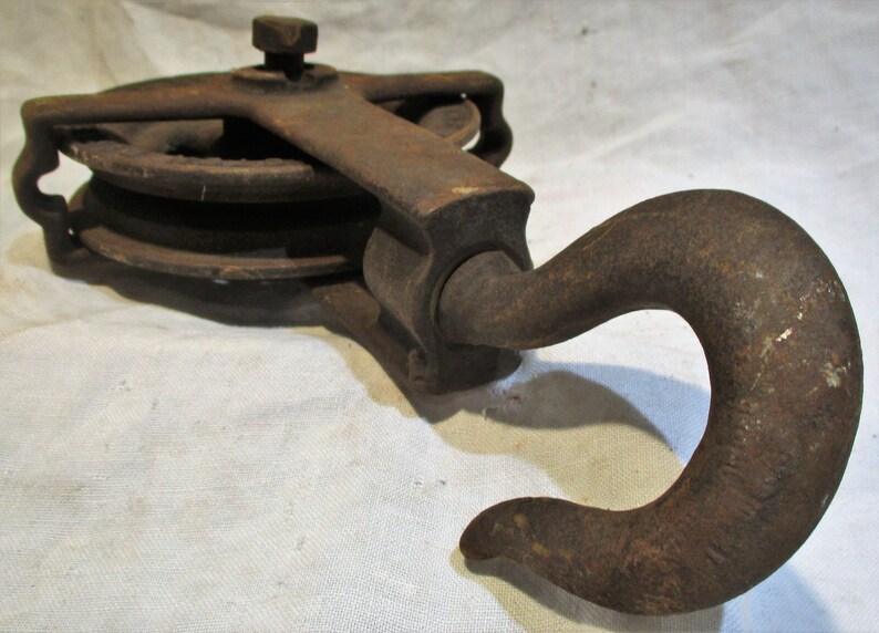 May include: A rusty cast iron pulley with a hook. The pulley has two wheels and a handle. The hook is bent at a 90-degree angle.