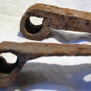 May include: Two rusty metal hinges with a circular hole in the center of each. The hinges are lying on a white fabric surface.