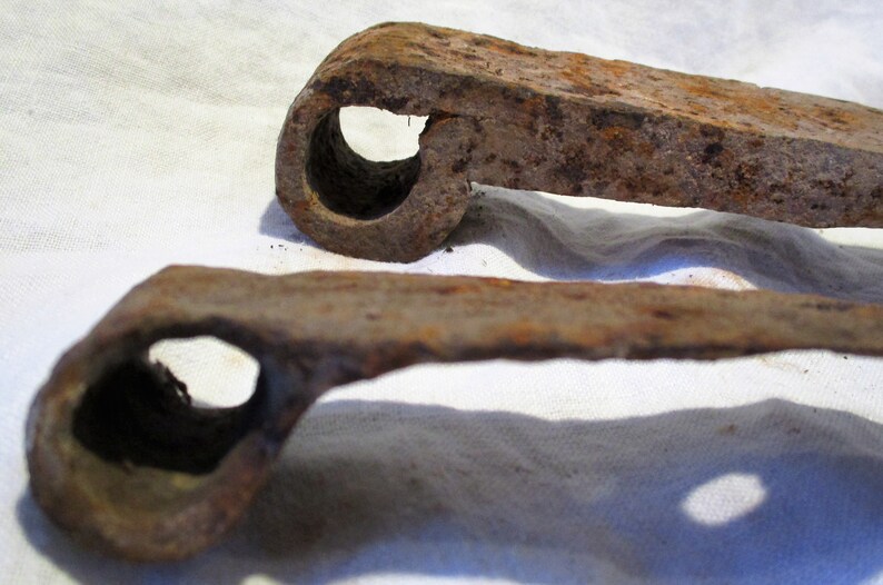 May include: Two rusty metal hinges with a circular hole in the center of each. The hinges are lying on a white fabric surface.