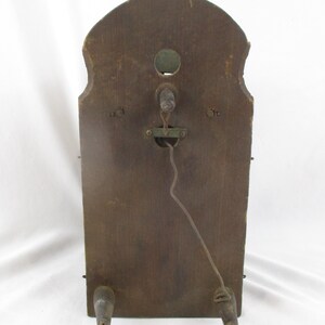 May include: A vintage wooden wall clock back with a dark brown finish. The clock has a curved top and two wooden feet. A metal mechanism and a wire are visible in the center. The clock's aged appearance suggests it is an antique.