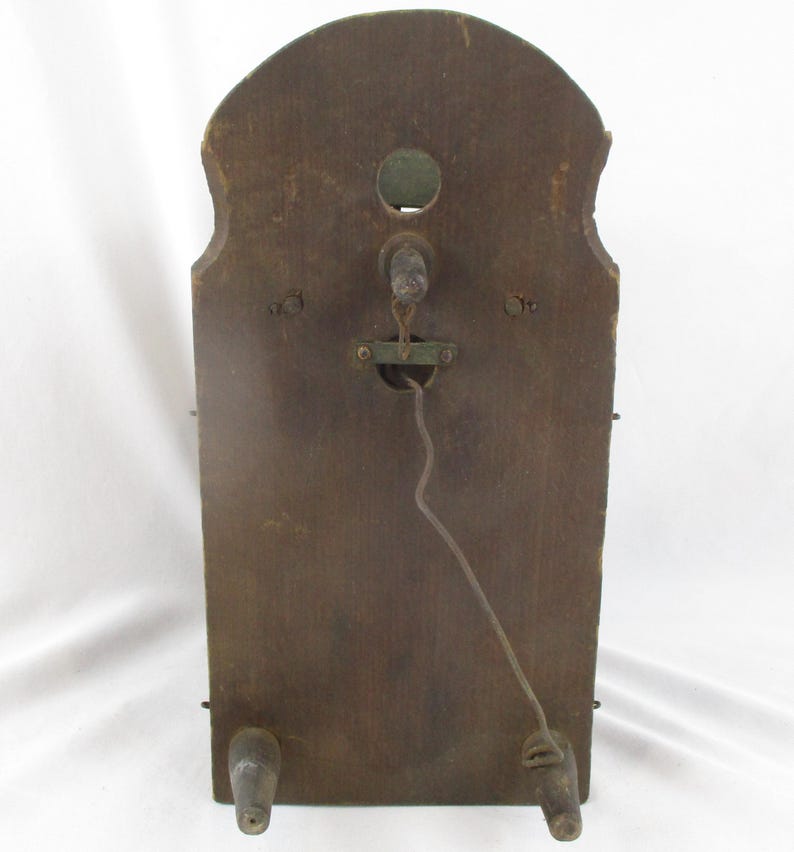 May include: A vintage wooden wall clock back with a dark brown finish. The clock has a curved top and two wooden feet. A metal mechanism and a wire are visible in the center. The clock's aged appearance suggests it is an antique.