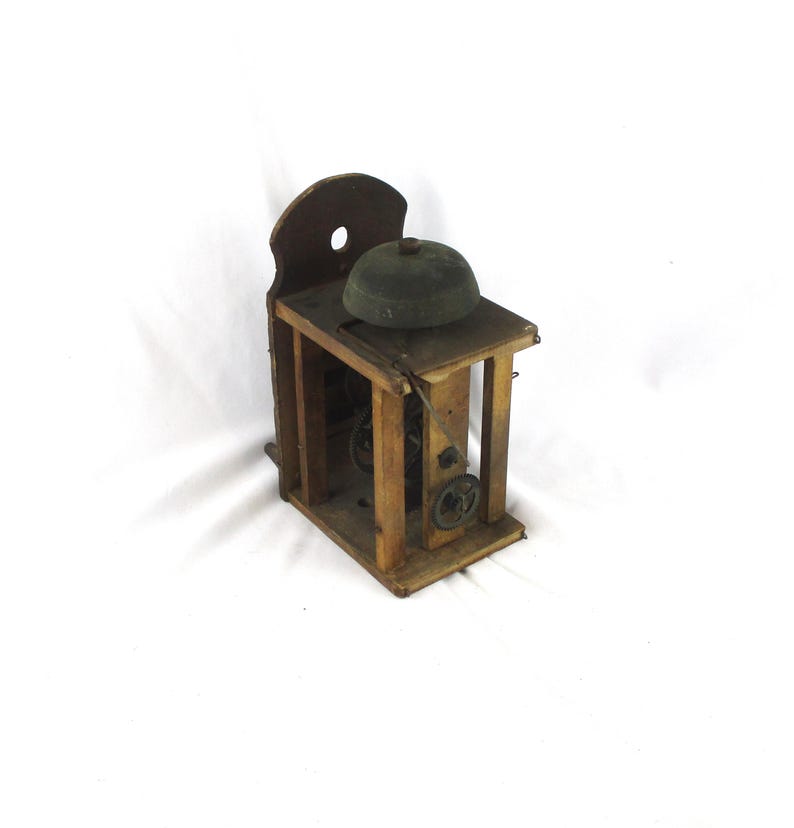May include: An antique wooden bell with a dark green bell and a wooden frame. The bell is mounted on a wooden platform with visible gears and a wooden backboard. The overall design is rustic and vintage.