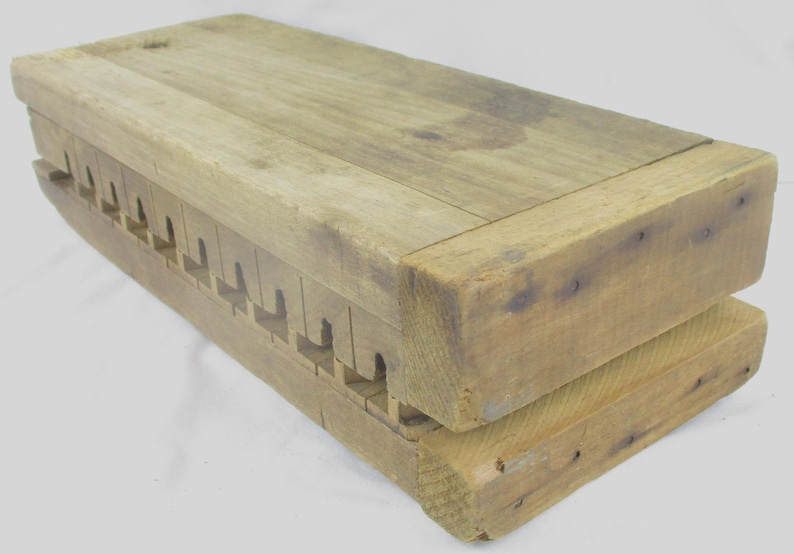 May include: A rectangular wooden mold featuring a natural light brown finish with visible wood grain. The object consists of two stacked blocks with a series of evenly spaced, notched cutouts along the side, joined by small metal nails.