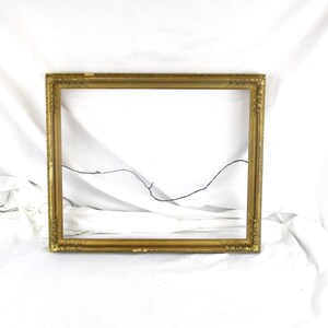May include: A rectangular picture frame with a metallic gold finish and decorative carved detailing along the edges. The frame features a thin, dark wire stretched horizontally across the center opening for hanging purposes.