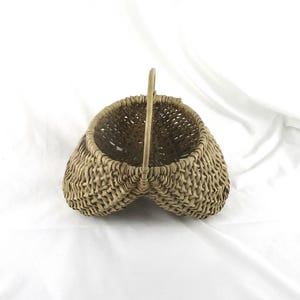 May include: A woven basket with a handle, in a heart shape. The basket is made of light brown wicker and has a sturdy handle. The basket is empty and sits on a white surface.