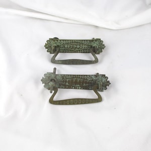 May include: Two antique bronze drawer pulls with ornate floral designs. The pulls have a dark green patina and curved handles. The decorative hardware is set against a white background.