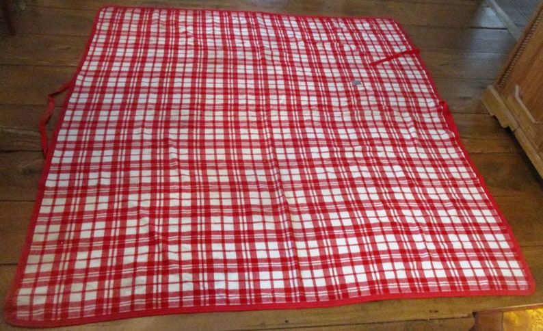 May include: Red and white checkered picnic blanket with a red border. The blanket is folded in half and has a strap on one side.