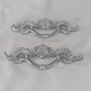 May include: Two silver-toned decorative drawer pulls with a keyhole design. The pulls have a scrolled, ornate design.