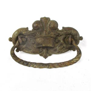 May include: A brass-toned metal drawer pull featuring a central crown motif flanked by two dolphin-like figures. The hardware includes a curved, twisted-rope style drop handle attached to a decorative backplate with a weathered, aged finish.