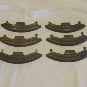 May include: Six antique brass drawer pulls with a curved, rounded shape and a raised, textured design. The pulls have a simple, classic style.