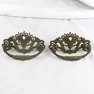 May include: Two antique brass drawer pulls with ornate, heart-shaped designs. The pulls have a curved handle and decorative detailing, perfect for vintage furniture restoration projects. The brass has a warm, aged patina.
