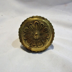 May include: A gold-toned metal knob with a decorative floral design. The knob has a raised center and a scalloped edge.