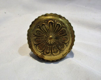 May include: A gold-toned metal knob with a decorative floral design. The knob has a raised center and a scalloped edge.