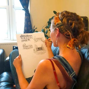May include: A person sketching architectural drawings on white paper with a pencil. The drawings depict building facades. The person is wearing denim overalls and a red top. A patterned scarf is wrapped around the person's head.
