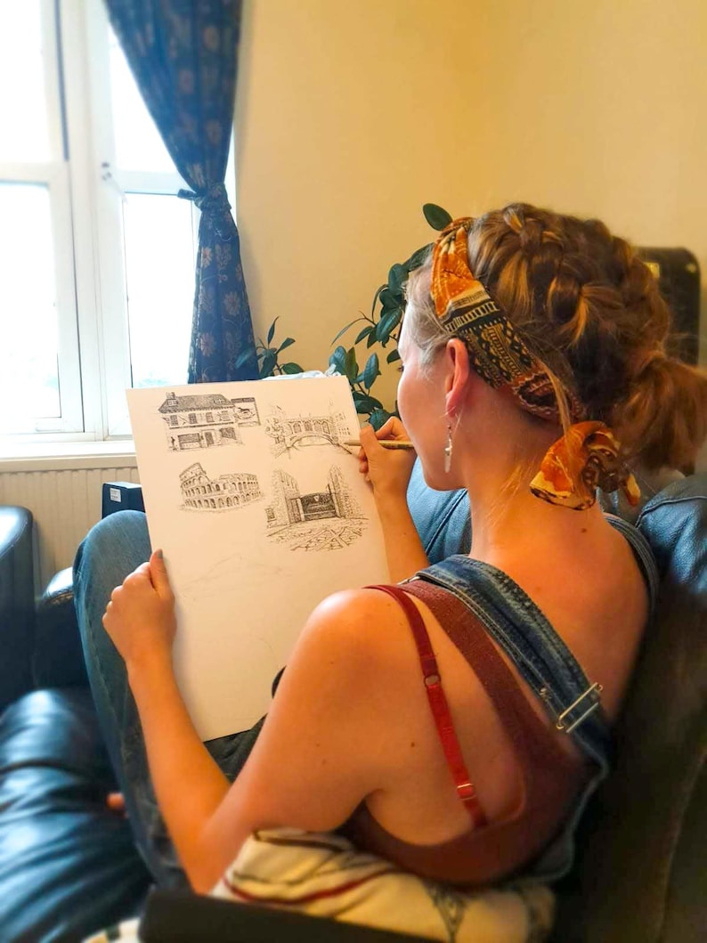 May include: A person sketching architectural drawings on white paper with a pencil. The drawings depict building facades. The person is wearing denim overalls and a red top. A patterned scarf is wrapped around the person's head.