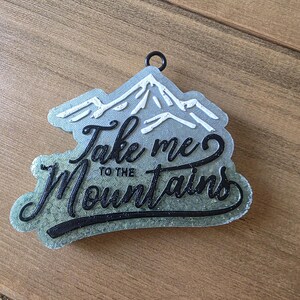 May include: A mountain-shaped ornament with the text "Take me to the Mountains" in black script. The mountains are white, and the base is a glittery green. A black ring is at the top for hanging.