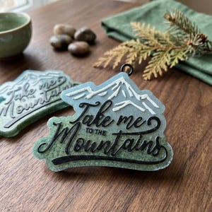 May include: A mountain-shaped ornament with the text "Take me to the Mountains" in black script. The ornament has a blue and green color scheme, with a mountain design in white. A black metal loop is attached for hanging.