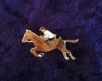 Puede incluir: Un pin de tono dorado con un caballo marrón y un jinete con camisa blanca y pantalón azul. El caballo está saltando una valla.