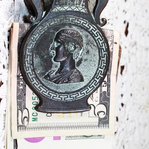 May include: Antique bronze-toned money clip featuring a classical profile within a circular frame. The clip is shaped like a vase with handles and holds several U.S. five-dollar bills. The background is a white, textured surface.