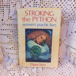 May include: A vintage book titled "STROKING the PYTHON women's psychic lives" by Diane Stein. The cover features an illustration of a woman with red hair, a blue garment, and a green orb against a starry background. The book is set against a white lace background.