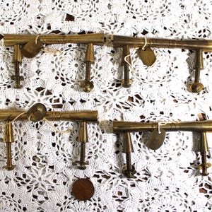 May include: Four antique brass furniture clamps with threaded rods and small metal tags. The clamps are arranged on a white lace doily. A penny is visible near the bottom.