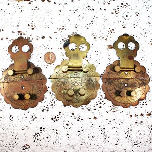 May include: Three antique brass latches with decorative floral and landscape engravings. Each latch features a unique design with a central circular element and a decorative top. The latches are set against a white, patterned background.