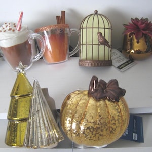 Puede incluir: Surtido de portavelas decorativos de vidrio en varias formas y colores, incluyendo una calabaza, un árbol de Navidad, una jaula de pájaros, una taza y una taza de chocolate caliente. Los portavelas están todos encendidos y tienen un brillo cálido.