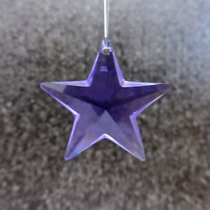 May include: A faceted, purple star-shaped ornament hangs suspended. The star has five points and is made of a translucent material, reflecting light. The background is a textured, gray surface. This decorative item could be used as a suncatcher or Christmas ornament.