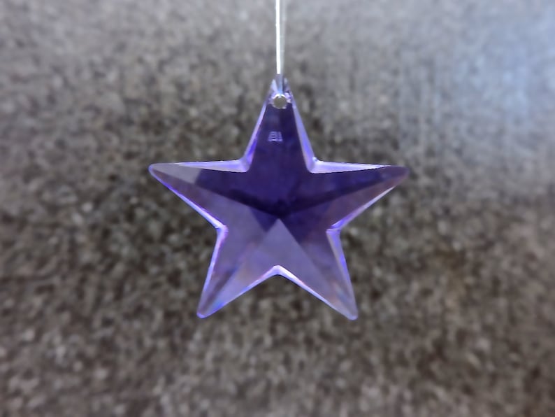 May include: A hanging, purple star-shaped ornament with a faceted design. The star is made of a translucent material, reflecting light. The background is a textured, grey surface. This decorative item could be used as a suncatcher or Christmas ornament.