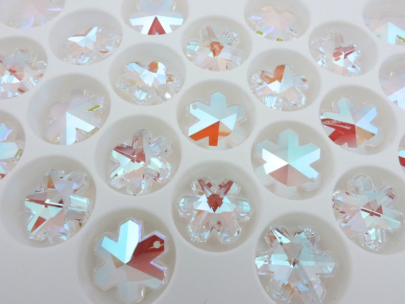 Peut inclure: Gros plan sur des cristaux transparents iris&eacute;s en forme de flocons de neige dispos&eacute;s dans un plateau blanc. Les cristaux ont de multiples facettes qui r&eacute;fl&eacute;chissent la lumi&egrave;re, cr&eacute;ant un effet arc-en-ciel.