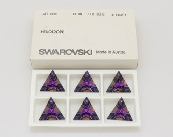 Puede incluir: Una caja de cristales Swarovski, etiquetada como "HELIOTROPE", con seis colgantes triangulares. Cada colgante es de un púrpura vibrante y mide 3 cm. La caja blanca tiene texto negro y el logotipo de Swarovski.