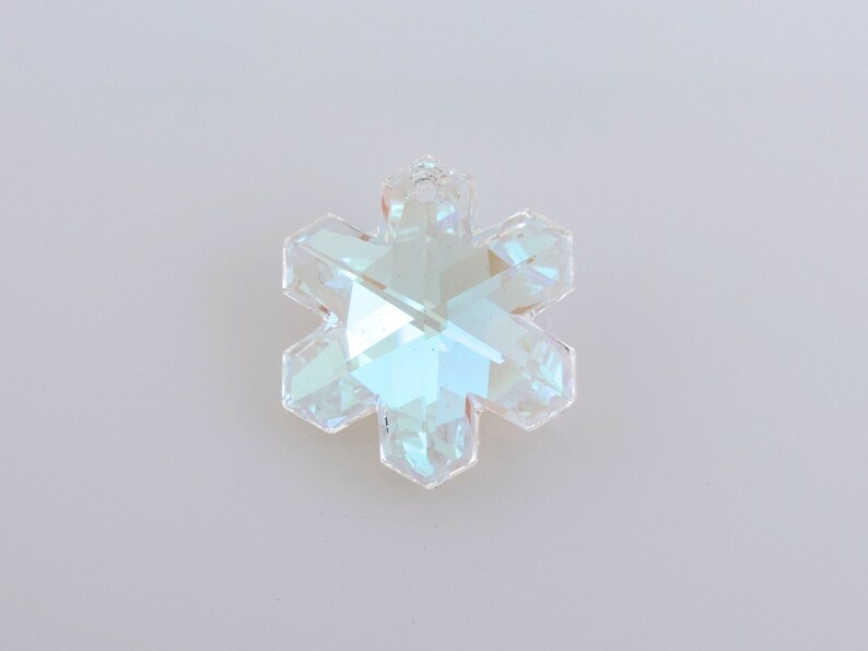 Peut inclure: Pendentif en cristal clair en forme de flocon de neige facett&eacute;. Le pendentif a six pointes et refl&egrave;te un arc-en-ciel de couleurs. Le cristal est suspendu sur un fond blanc uni. Id&eacute;al pour la fabrication de bijoux ou la d&eacute;coration de vacances.