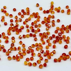 May include: A collection of small, faceted beads in shades of orange and yellow. The beads are scattered on a white surface, reflecting light. These beads are suitable for jewellery making and other craft projects.