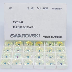 May include: A white box containing Swarovski crystals, labeled "Crystal Aurore Boreale." The crystals are iridescent, with a diameter of 18 mm. The box is marked "Made in Austria."