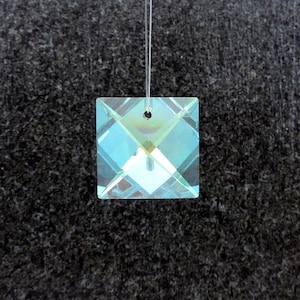 May include: Square crystal prism pendant with iridescent color reflections. The pendant is suspended by a thin, clear string. The crystal has a hole at the top for hanging. The background is a dark, textured surface.