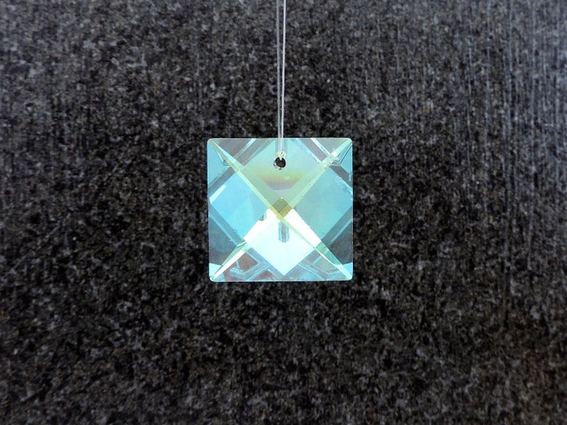 Puede incluir: Colgante de prisma de cristal cuadrado con reflejos de color iridiscente. El colgante est&aacute; suspendido por un hilo fino y transparente. El cristal tiene un agujero en la parte superior para colgarlo. El fondo es una superficie oscura y texturizada.