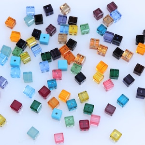 May include: Assortment of colorful square glass beads in various shades of blue, green, red, orange, yellow, and purple. The beads are scattered on a white surface.