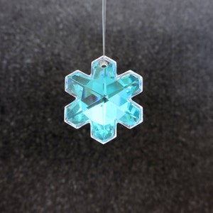 Peut inclure: Ornement en forme de flocon de neige clair et facett&eacute;, de couleur turquoise. Le cristal &agrave; six pointes est suspendu par un fil fin et transparent. L'ornement refl&egrave;te la lumi&egrave;re, cr&eacute;ant un effet scintillant. Le fond est une surface sombre et textur&eacute;e.