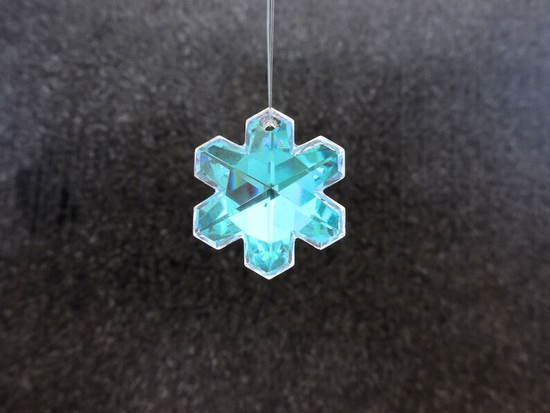 Peut inclure: Ornement en forme de flocon de neige clair et facett&eacute;, de couleur turquoise. Le cristal &agrave; six pointes est suspendu par un fil fin et transparent. L'ornement refl&egrave;te la lumi&egrave;re, cr&eacute;ant un effet scintillant. Le fond est une surface sombre et textur&eacute;e.