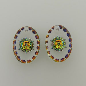 May include: A pair of oval glass pendants featuring a sun design. The sun has an iridescent finish with a silver frame. The edges of the pendants are decorated with small, colourful dots. Each has a small hole.