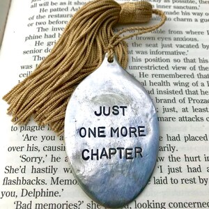 May include: A silver bookmark with a tassel. The bookmark is shaped like a spoon and has the text "Just One More Chapter" engraved on it.