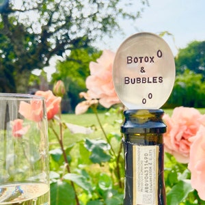 May include: Silver bottle stopper with the text "BOTOX O & BUBBLES 00" on a champagne bottle in a garden setting.