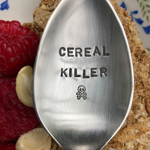 Op de afbeelding: Een zilveren lepel met de woorden "Cereal Killer" gegraveerd, samen met een schedel en gekruiste beenderen.